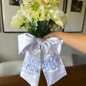 May include: A bouquet of cream and white artificial flowers tied with a large white satin ribbon. The ribbon features blue embroidery with the text HOCO 2026 on one tail and a decorative three-letter monogram on the other tail.