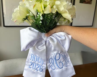 May include: A bouquet of cream and white artificial flowers tied with a large white satin ribbon. The ribbon features blue embroidery with the text HOCO 2026 on one tail and a decorative three-letter monogram on the other tail.