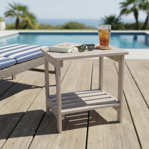 May include: A light gray wooden side table with a slatted bottom shelf. A book, sunglasses, and a glass of iced tea sit on top. The table is on a wooden deck near a pool.