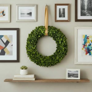 May include: A vibrant green wreath made of boxwood, suspended by a gold ribbon against a neutral wall. The wreath is the focal point, surrounded by framed artwork and a wooden shelf with decor, creating a welcoming home decor scene.
