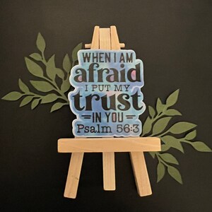 May include: A sticker with the text "WHEN I AM AFRAID I PUT MY TRUST IN YOU Psalm 56:3" is displayed on a small wooden easel. The sticker has a watercolor effect with blue, purple, and white hues. Green leaf cutouts are on the sides.