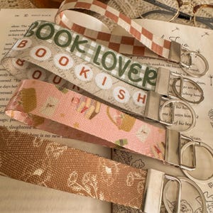 May include: Several fabric keyrings with silver-coloured metal fixings. The keyrings display patterns such as a checkerboard, the text "BOOK LOVER", and floral and book-themed designs. The keyrings are arranged on an open book.