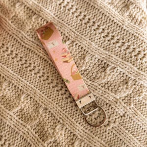 May include: A pink fabric keychain wristlet featuring a pattern of baskets, cakes, and other items. It has a silver-tone metal clasp and a key ring. The wristlet is placed on a cream-coloured knitted surface.