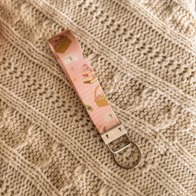 May include: A pink fabric keychain wristlet featuring a pattern of baskets, cakes, and other items. It has a silver-tone metal clasp and a key ring. The wristlet is placed on a cream-coloured knitted surface.