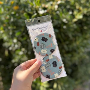 May include: Two round car coasters with a blue background and a design of dice, cherries, and playing cards. The coasters are packaged in a clear plastic bag with a white card that says "Car Coasters Handmade with love".
