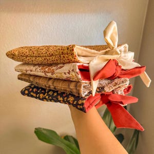 Könnte beinhalten: Ein Stapel gesteppter Stoffbeutel in verschiedenen floralen und gemusterten Designs. Die Beutel sind mit cremefarbenen und roten Schleifen gebunden. Die Stoffe weisen Farben wie Braun, Creme, Rot und Schwarz auf.
