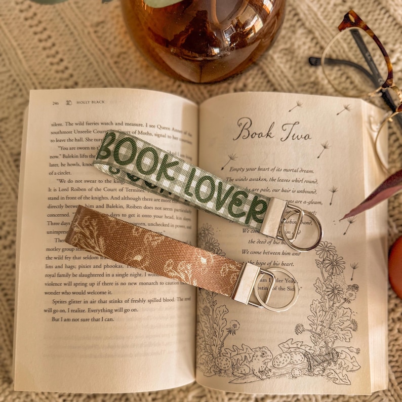 May include: Two keychains lie on an open book. One is green and white with "BOOK LOVER" in green. The other is brown with a floral pattern. The book is open to a page with text and the words "Book Two".