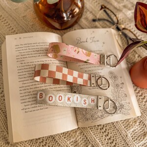 May include: Three fabric keyrings with metal fixings lie on an open book. One is pink with a floral pattern, another is a peach and white checkerboard, and the third reads "BOOKISH" in lettered blocks.