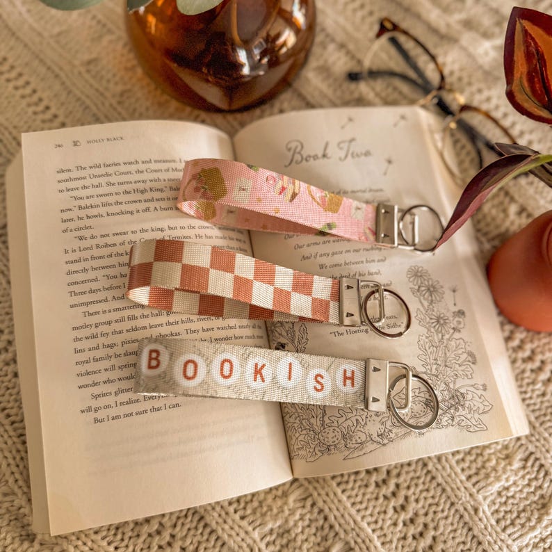 May include: Three fabric keyrings with metal fixings lie on an open book. One is pink with a floral pattern, another is a peach and white checkerboard, and the third reads "BOOKISH" in lettered blocks.