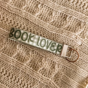 May include: A fabric keychain with the words "BOOK LOVER" printed in green on a white and green checkered pattern. The keychain has a silver metal clasp and a bronze-coloured key ring. The background is a beige knitted fabric.