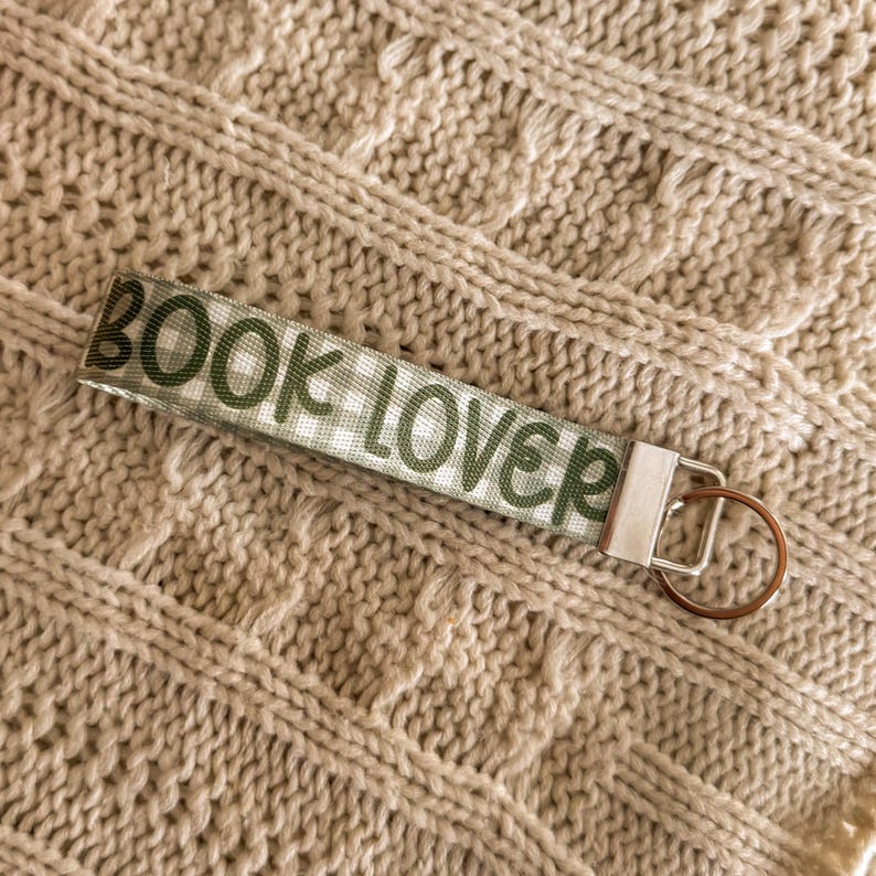 May include: A fabric keychain with the words "BOOK LOVER" printed in green on a white and green checkered pattern. The keychain has a silver metal clasp and a bronze-coloured key ring. The background is a beige knitted fabric.