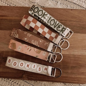 Pode incluir: Uma coleção de chaveiros de tecido com ferragens prateadas. Um chaveiro diz "BOOK LOVER" em letras verdes sobre um fundo xadrez cinza e branco. Outro diz "BOOKISH" em letras marrons. Outros chaveiros têm padrões florais e xadrez.
