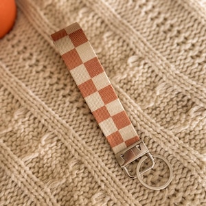 May include: A woven keychain wristlet with a cream and rust-orange checkerboard pattern. It features a silver-coloured metal clasp and key ring. The background is a cream-coloured knitted fabric.