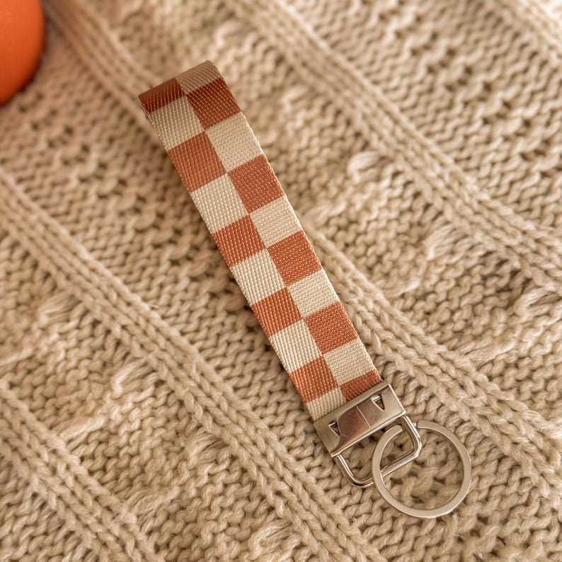 May include: A woven keychain wristlet with a cream and rust-orange checkerboard pattern. It features a silver-coloured metal clasp and key ring. The background is a cream-coloured knitted fabric.