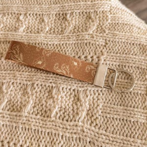 May include: A brown fabric keychain wristlet with a white floral pattern. The wristlet has a silver-coloured metal clasp and a split ring for keys. The background is a cream-coloured knitted fabric.