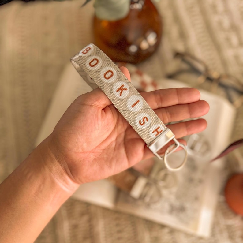 May include: A beige fabric keychain with the word "BOOKISH" in red and white letters. The keychain has a silver metal clasp and ring. The background includes a book, glasses, and a decorative vase.