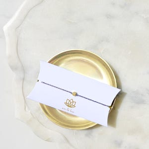 May include: A thin purple string bracelet featuring a small gold-colored bead, wrapped around a white rectangular card with curved edges. The card displays a gold lotus flower icon and the text emi & koa printed in a small, simple font.