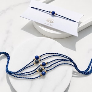 May include: A set of dark blue cord bracelets featuring round lapis lazuli and grey stone beads accented by small gold-toned metal spheres. One bracelet is displayed on a white card printed with the text emi & koa and a lotus flower icon.