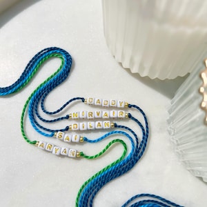 May include: Five handmade bracelets made of blue and green braided cord, each featuring white square beads with gold lettering spelling out the names DADDY, NIRVAIR, DILAN, SAI, and ARYAN, accented with small gold-coloured spacer beads.