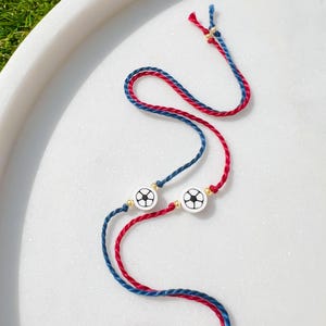 May include: Two bracelets with red and blue cords. Each bracelet features a white circular charm with a black football design. The bracelets are displayed on a white marble surface.