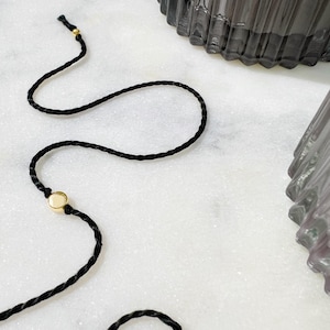 May include: A thin black braided cord bracelet featuring a single round gold-toned metal bead with a smooth surface. The cord is finished with a small gold-toned metal accent at the end, providing a simple and clean design for this wrist accessory.