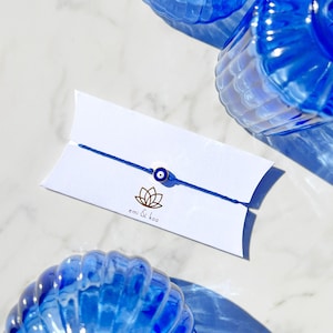 May include: A blue string bracelet with a gold-colored evil eye charm, presented on a white card with the logo "emi & koo". The bracelet is displayed with blue glass candle holders on a marble surface.