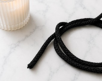 May include: A black braided cord arranged in a loose loop and a lit candle in a clear, vertically ribbed glass container. The cord features a textured, woven surface, while the candle emits a warm, steady flame from within the glass vessel.