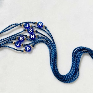 May include: A set of five blue silk cord bracelets, each featuring a single round blue bead with a white letter. The letters displayed are J, R, S, H, B, J, and A. Small metallic gold-toned spacer beads are positioned on either side of each letter bead.