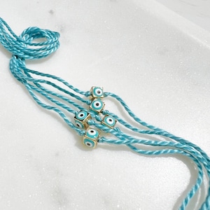 May include: Turquoise string bracelets with gold-colored cube beads, each featuring a blue and white evil eye design. The bracelets are knotted at the top and rest on a white surface.
