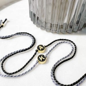 Könnte beinhalten: Zwei Armbänder aus gedrehter schwarzer und grauer Kordel, jeweils mit einem runden goldfarbenen Anhänger mit schwarzer Mitte. Ein Anhänger zeigt den Buchstaben A in Gold, der andere den Buchstaben K in Gold, beide mit glänzender Oberfläche.