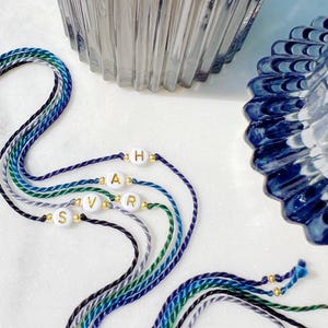 May include: Four braided cord bracelets in shades of blue, green, and grey. Each bracelet features a single white round bead with a gold letter, spelling out the name SARAH, accented by small gold-toned metallic spacer beads on either side.