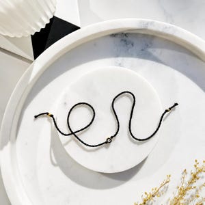 Peut inclure: Un bracelet en cordon noir fin doté d'une petite breloque œil porte-bonheur centrée avec un motif circulaire blanc, bleu et noir. Le bracelet comprend deux minuscules perles métalliques dorées positionnées près des extrémités du cordon.