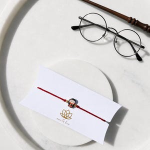 Peut inclure: Un bracelet en fil rouge fin avec une petite breloque colorée représentant un personnage aux cheveux noirs portant des lunettes. Le bracelet est présenté sur une carte blanche avec un logo lotus doré et le texte emi & koa, près de lunettes et d'une baguette.