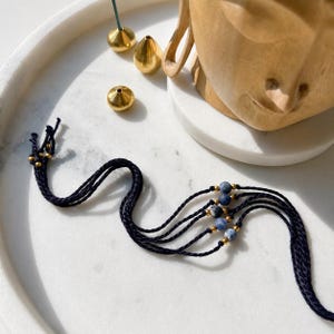 May include: A multi-strand bracelet featuring dark navy blue cord, small gold-toned metallic beads, and round blue and grey marbled stone beads. The bracelet consists of four separate strands that are joined together at the ends with loose cord.