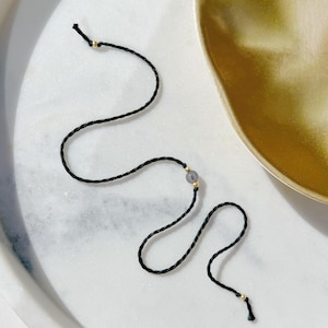 Könnte beinhalten: Ein zartes Armband aus einer dünnen, gedrehten schwarzen Kordel. Das Design umfasst eine einzelne kleine, durchscheinende graue Perle, die zwischen zwei winzigen goldfarbenen Metallabstandshaltern sitzt, sowie weitere goldfarbene Perlen an den Enden.