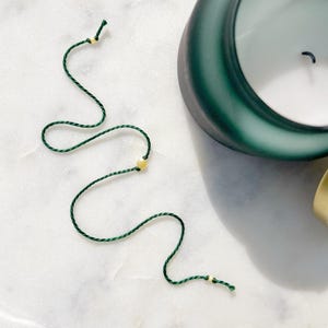 May include: A thin, dark green cord bracelet featuring three small, round, metallic gold-toned beads spaced along the length of the string. The cord is twisted and flexible, with knotted ends securing the beads in place along the delicate strand.