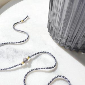 May include: A thin, twisted grey cord bracelet featuring a single translucent grey bead flanked by two small, shiny gold-toned metallic spheres. The cord is flexible and lightweight, with a simple design that highlights the contrast between the grey tones.
