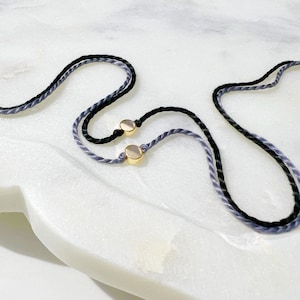 May include: Two thin, braided cords, one in black and one in a muted lavender-grey, each featuring a small, polished gold-tone cylindrical bead. The cords are arranged in a curved, overlapping pattern, highlighting the contrast between the dark and light fibers.