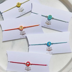 Könnte beinhalten: Eine Sammlung farbenfroher Armbänder mit runden, illustrierten Anhängern. Jedes Armband hat eine andersfarbige Kordel und einen Anhänger, präsentiert auf weißen Karten mit dem Text "emi & koa" und einem Lotusblumen-Logo.