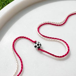 Puede incluir: Una pulsera con un pequeño dije de balón de fútbol en blanco y negro. La pulsera tiene un cordón trenzado rojo y blanco. La pulsera se muestra sobre una superficie blanca.
