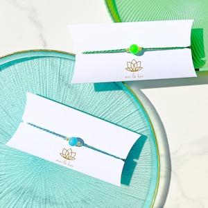 Può includere: Due braccialetti con cordino su cartoncini bianchi con il testo emi & koo e un logo a forma di loto. Un braccialetto presenta un cordino verde brillante con una perla verde neon, l'altro un cordino verde acqua con una perla azzurra, entrambi con perline dorate.