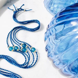 May include: A set of four dark blue braided cord necklaces featuring small, round, frosted teal beads and tiny gold-toned metallic spacers. The cords are arranged in a loose, flowing pattern alongside a large, ribbed, translucent blue glass object.