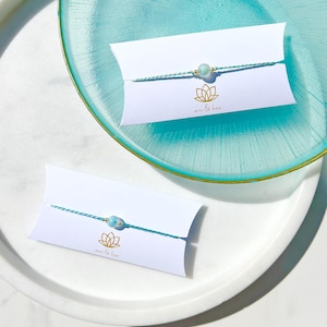 May include: Two bracelets featuring a single light blue bead on a thin, teal and white twisted cord. Each bracelet is attached to a white rectangular card with a gold lotus flower logo and the text emi & hoa printed in small gold letters.