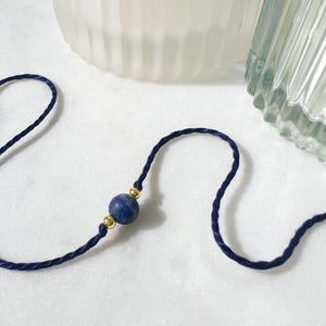 May include: A dark blue twisted cord bracelet featuring a single round blue stone bead centred between two small, polished gold-tone metal beads. The cord is thin and flexible, creating a simple, minimalist design with a smooth, uniform texture.
