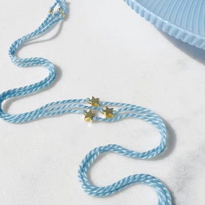 May include: A light blue braided cord bracelet featuring three small gold-toned star charms attached to the centre. The cord is constructed from multiple strands of twisted light blue thread, and the ends are finished with small gold-toned beads.