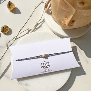 Könnte beinhalten: Ein minimalistisches Armband mit einer kleinen, runden, weiß-grauen Steinperle auf einer grauen Kordel. Goldfarbene Perlen akzentuieren die Kordel. Das Armband wird auf einer weißen Karte mit dem Text "emi & koa" und einem Lotusblumen-Logo präsentiert.