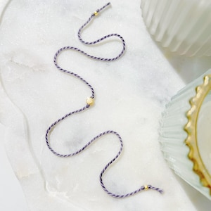 May include: A thin bracelet made of twisted purple and white cord, featuring three small, round, polished gold-coloured metal beads spaced along the length of the string. The cord is arranged in a loose, wavy, serpentine pattern on a white surface.