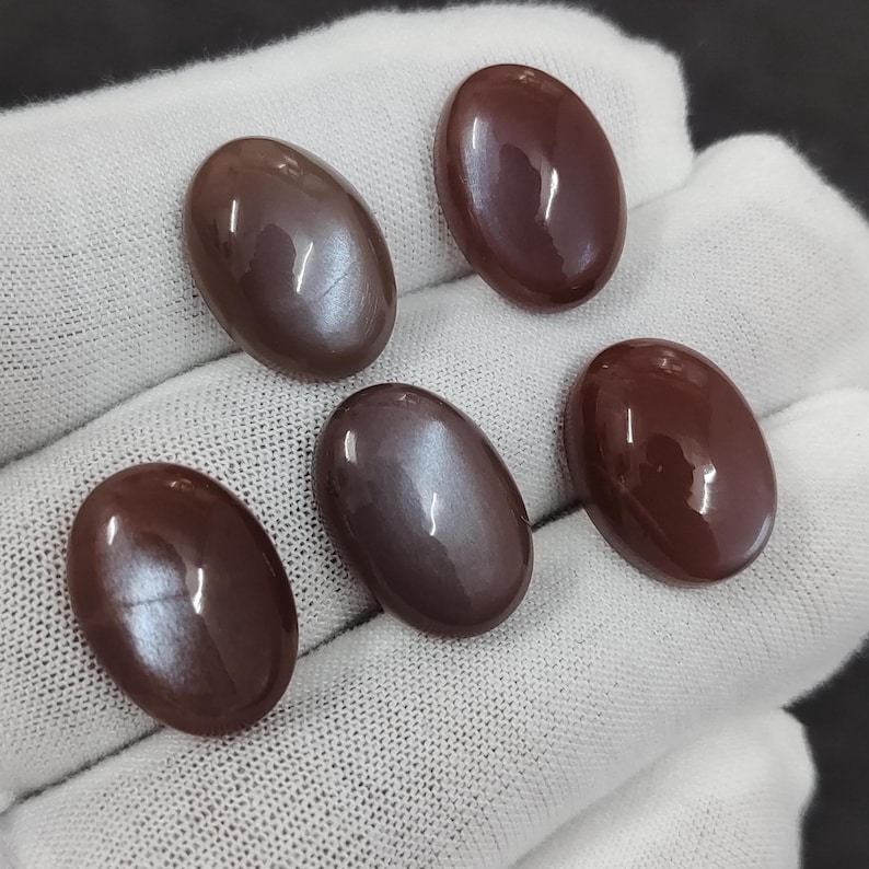 May include: Five oval-shaped gemstones with a brown and iridescent sheen. The stones are arranged in a row on a white surface.