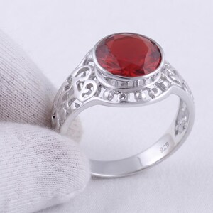 May include: A silver ring featuring a large, round, faceted red gemstone set in a bezel. The band displays an intricate, openwork filigree pattern along the sides and is stamped with the number 925 on the inner surface to indicate sterling silver.
