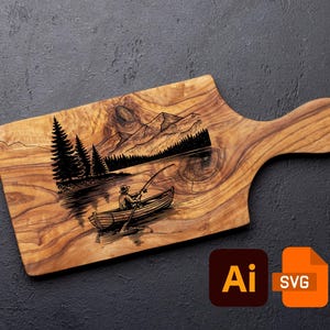 Könnte beinhalten: Holzschneidebrett mit Griff, mit einer gravierten Szene einer Person, die von einem Boot aus auf einem See fischt, mit Bergen und Bäumen im Hintergrund. Das Brett hat eine natürliche Holzmaserung.